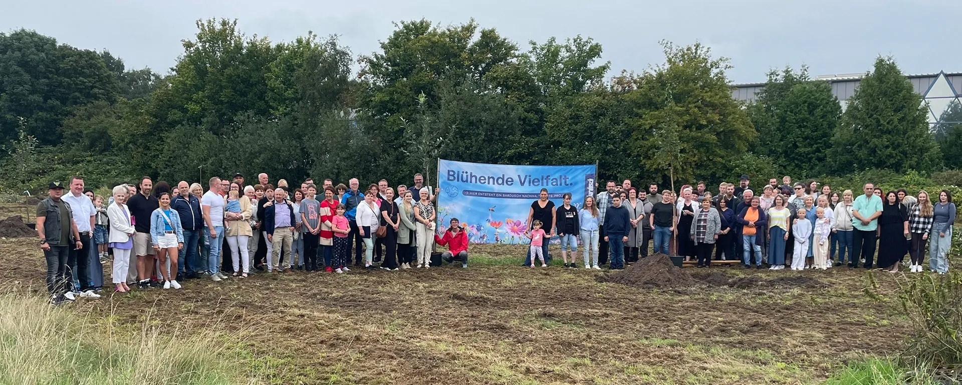 Eine große Gruppe von etwa hundert Bardusch-Mitarbeitern und ihren Familien steht für ein Gruppenfoto auf einem frisch bearbeiteten Feld mit Erdhügeln. Hinter ihnen hängt ein blaues Banner mit der Aufschrift „Blühende Vielfalt“.