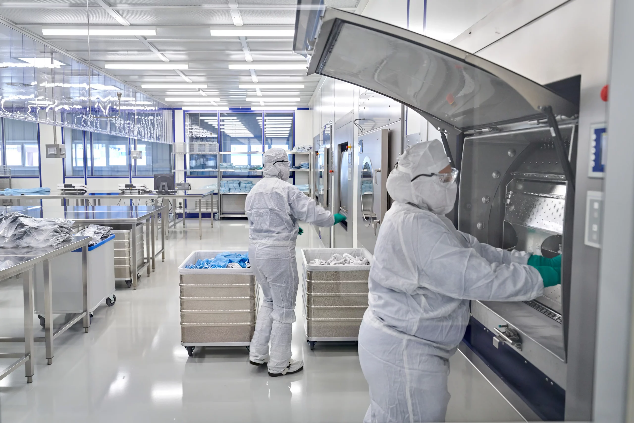 Zwei Mitarbeiter in weißen Reinraumanzügen und Schutzausrüstung (Goggles, Handschuhe) arbeiten in einer hellen, hochreinen Produktionsumgebung. Sie sind dabei, Textilien in großen, glänzenden Industriewaschmaschinen zu laden oder zu entnehmen.