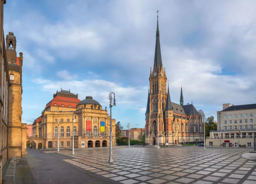 Stadtansicht des Opernhauses und der St.-Petri-Kirche in Chemnitz mit großem Platz im Vordergrund und heller Morgenstimmung.