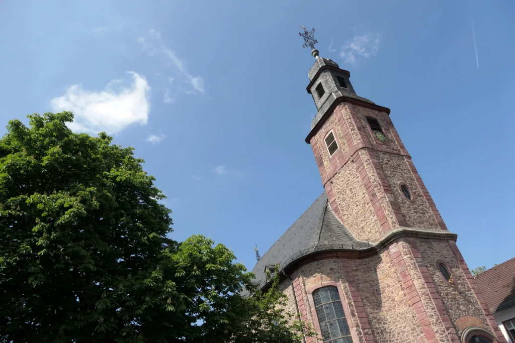 Schräg aufgenommene Ansicht einer historischen Kirche aus rotem Sandstein in Dreieich mit Turm, Schieferdach und umgebenden Bäumen unter blauem Himmel.