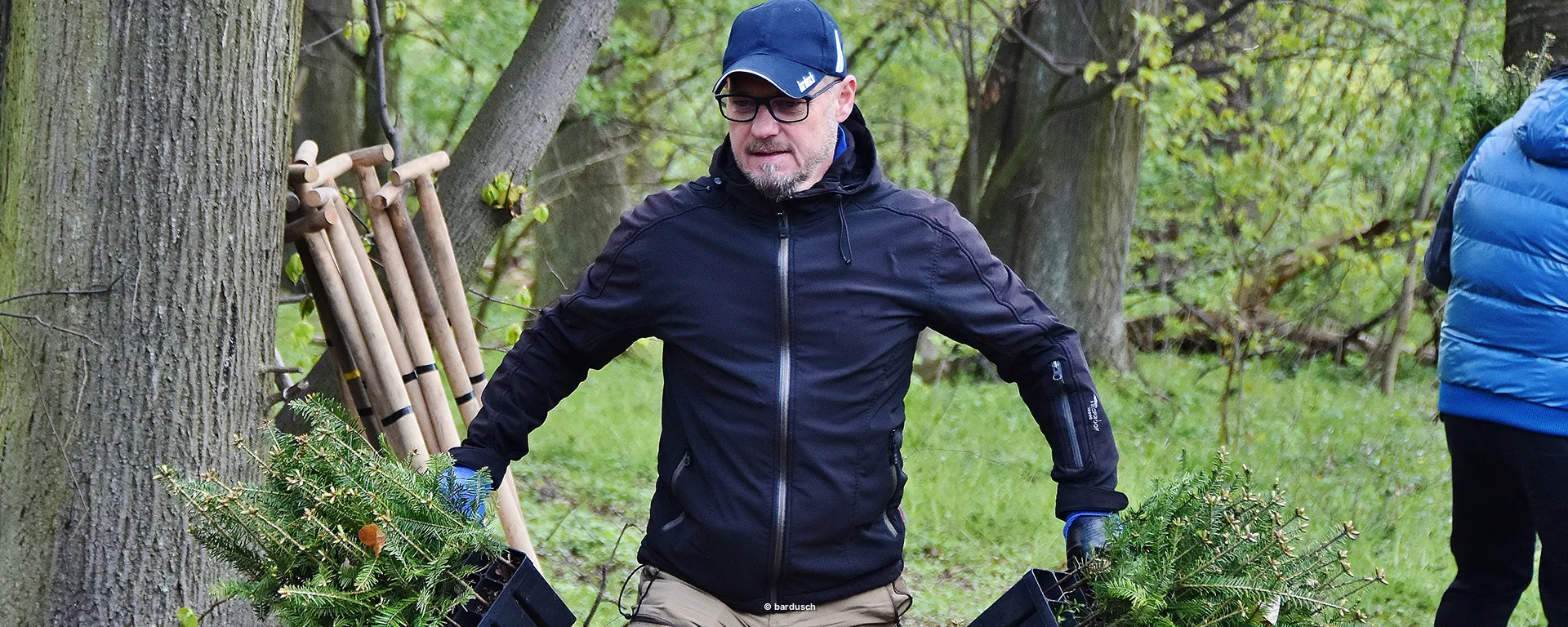 Ein Mann mit Mütze und blauen Handschuhen trägt in einem lichten Nadelwald zwei schwarze Kisten voller junger, grüner Baumsetzlinge.