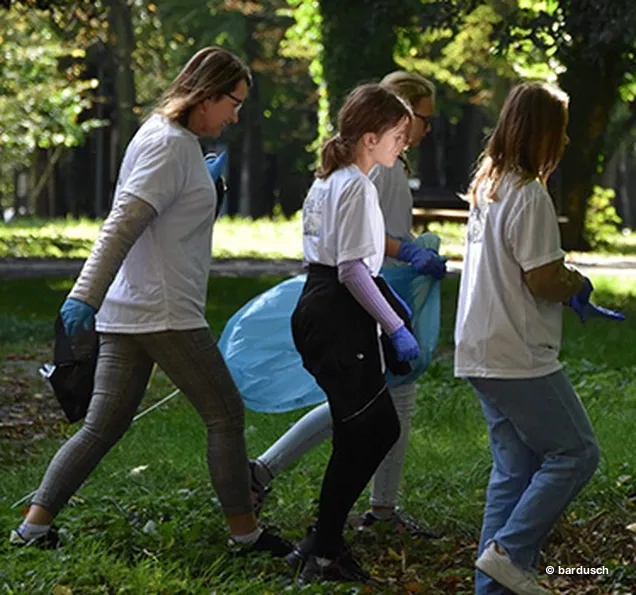 Drei Jugendliche gehen in weißen T-Shirts und dunklen Hosen auf einem Pfad und tragen gefüllte Abfallsäcke.