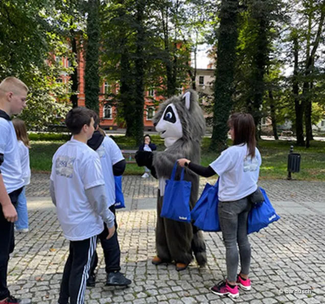Zwei Jugendliche und ein Erwachsener interagieren mit dem Waschbär-Maskottchen auf einem Waldweg.