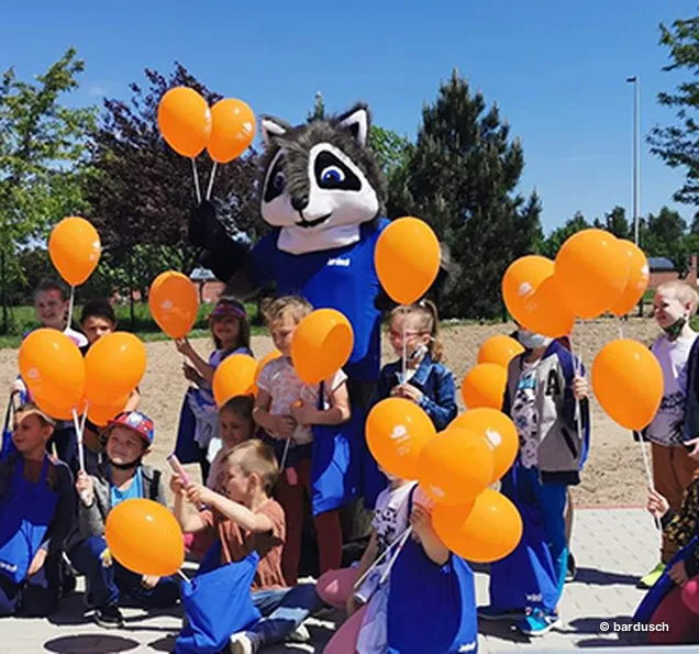 Ein Waschbär-Maskottchen spielt mit einem Tischtennisschläger an einer neuen Tischtennisplatte im Freien, umgeben von einer großen Gruppe jubelnder Kinder.