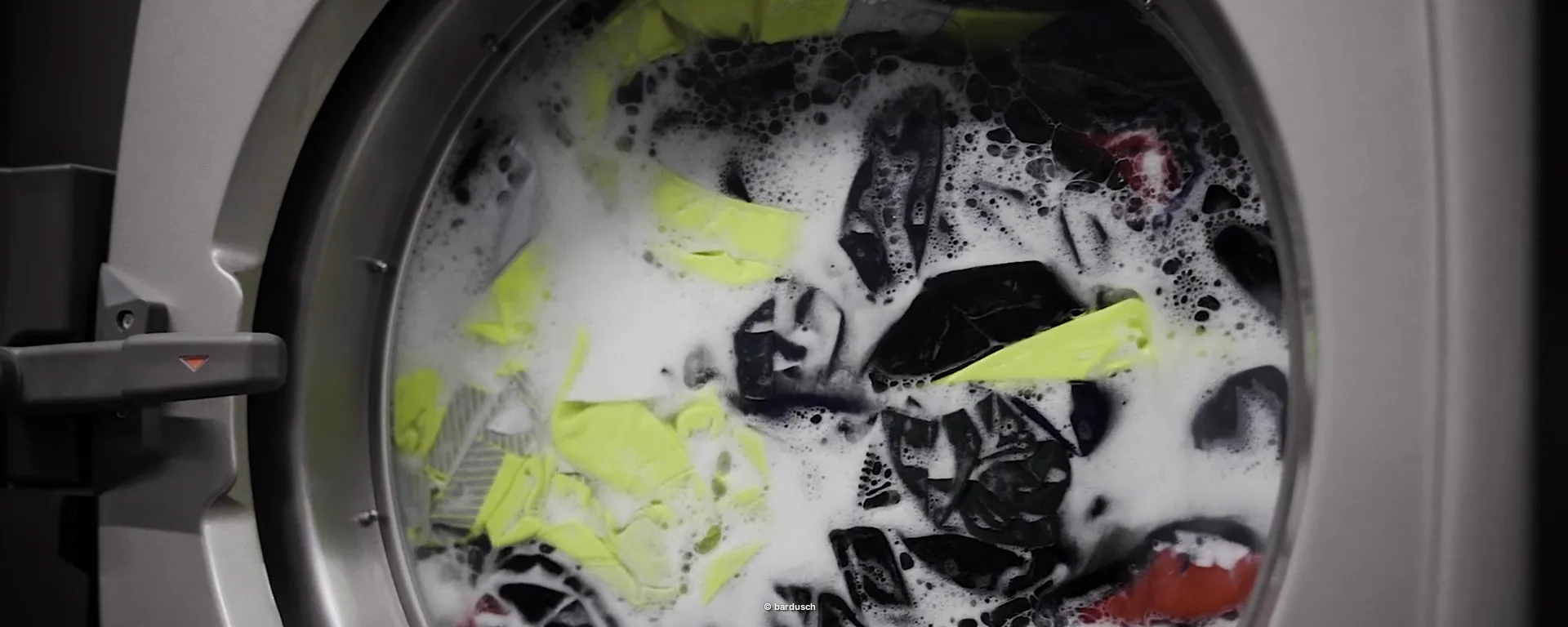 Nahaufnahme einer Waschmaschinentrommel während des Betriebs. Darin drehen sich nasse, schäumende Textilien, darunter dunkle und auffällig gelbe (Warnschutz-)Kleidungsstücke.