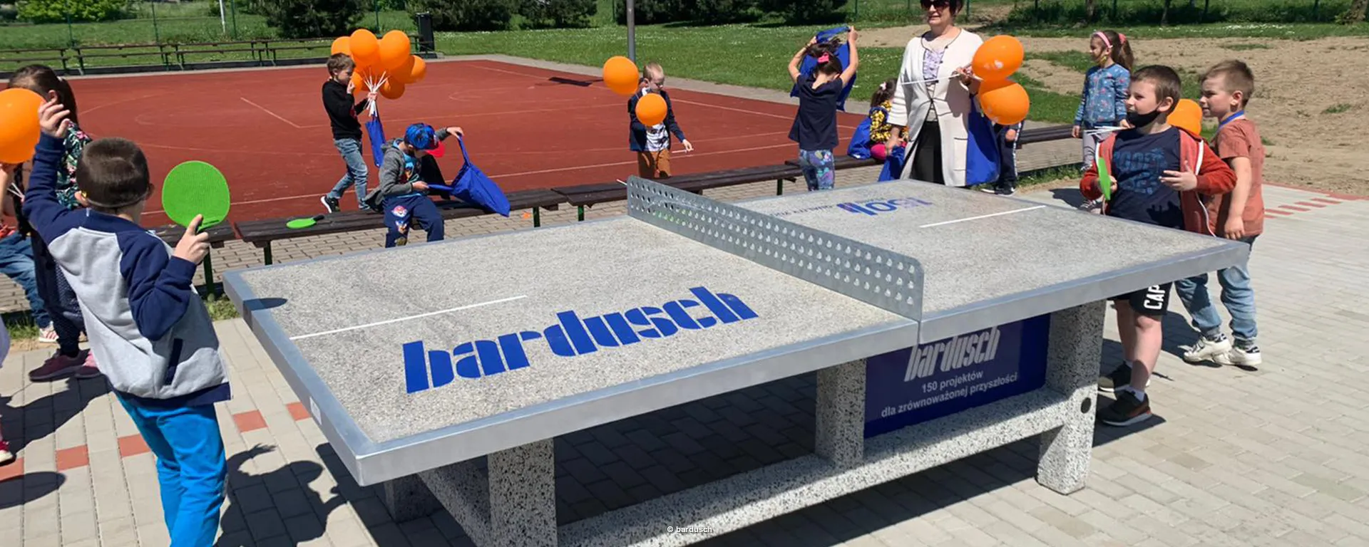Eine neue, robuste Outdoor-Tischtennisplatte mit dem blauen "bardusch"-Logo. Kinder stehen ringsherum, halten orangefarbene Ballons und spielen auf dem angrenzenden Sportplatz.