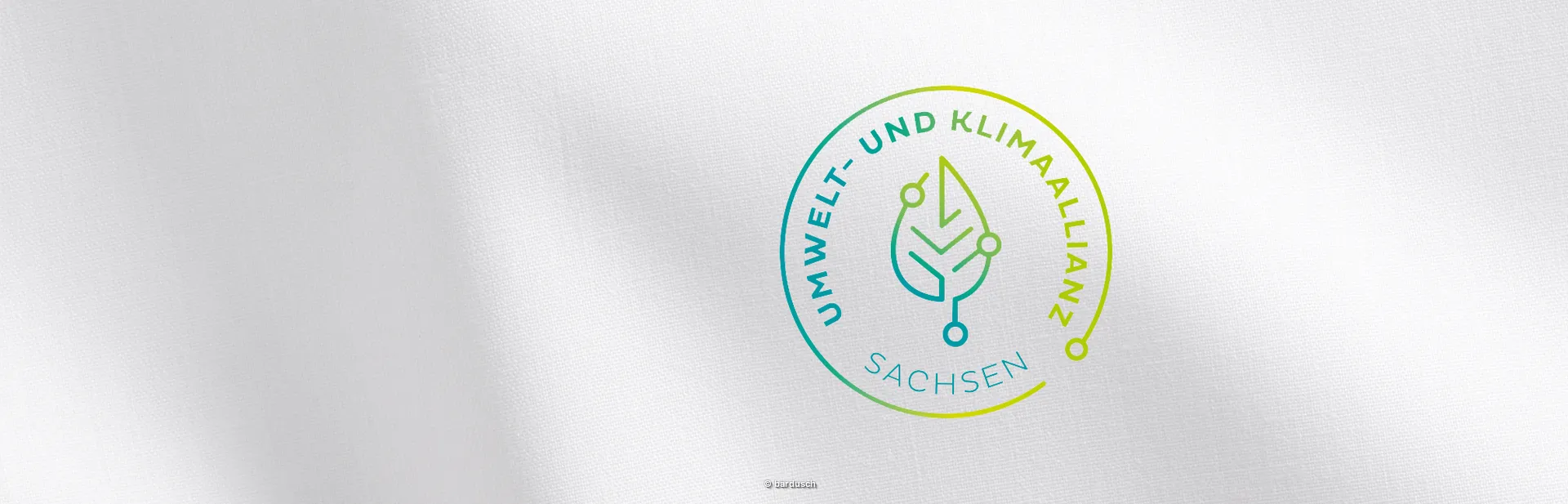 Das offizielle Logo der Umwelt- und Klimaallianz Sachsen. Es zeigt in einem runden Kranz mit grün-gelbem Farbverlauf die Worte "UMWELT- UND KLIMAALLIANZ SACHSEN" und in der Mitte ein stilisiertes Blatt mit technologischen Verbindungen.