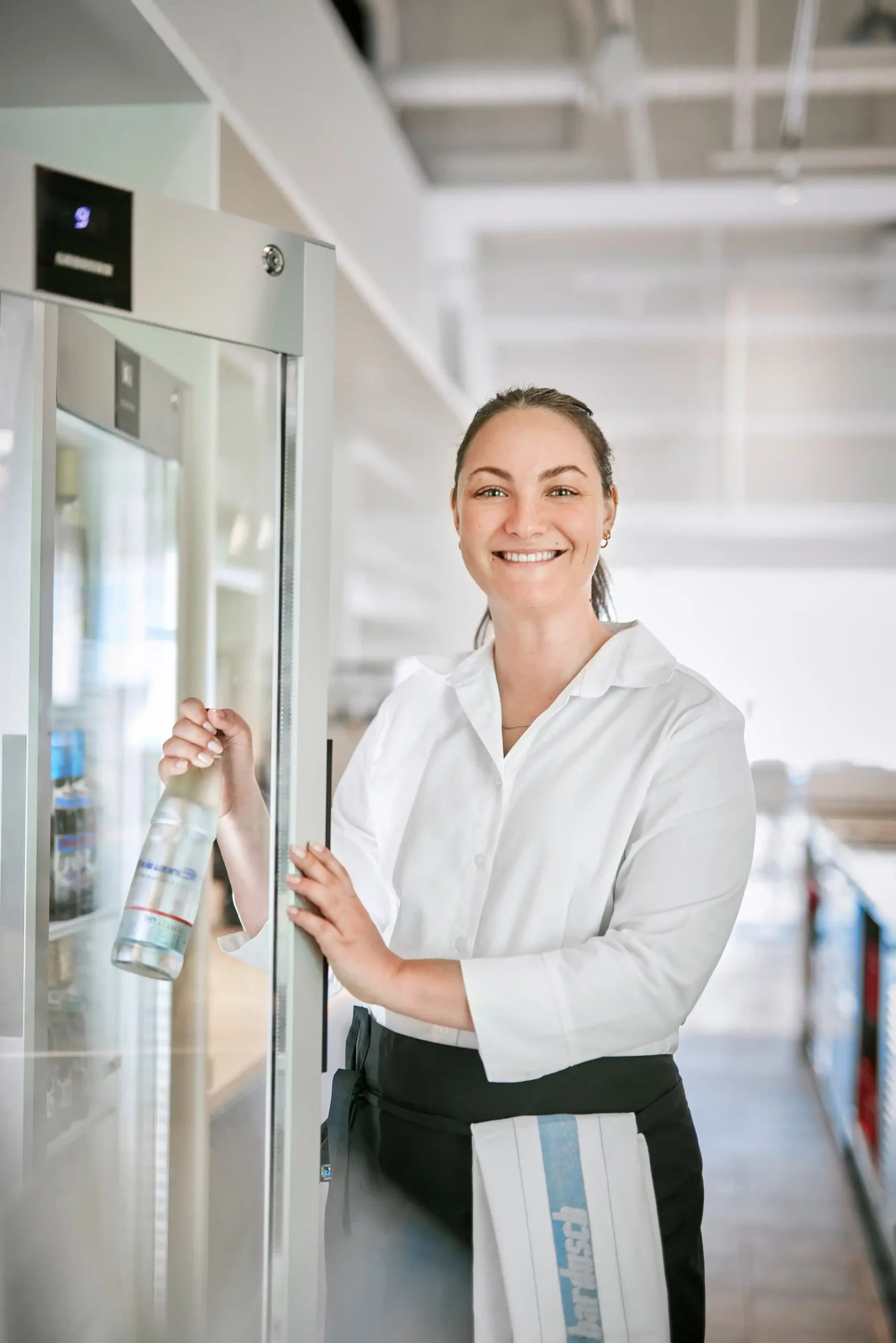 Servicekraft in weißer Bluse holt eine Wasserflasche aus einem Kühlschrank in einer Gastroküche