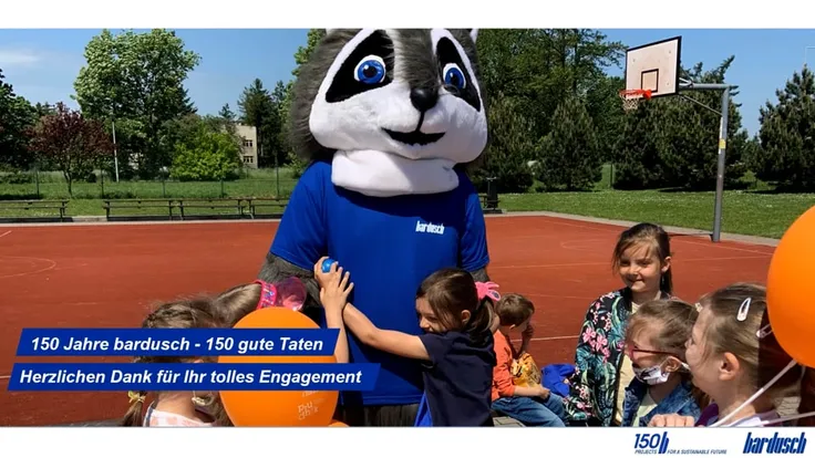 Eine Gruppe kleiner Kinder auf einem roten Sportplatz interagiert fröhlich mit einem großen Waschbär-Maskottchen in blauem T-Shirt. Im Vordergrund sind orangefarbene Ballons zu sehen, im Hintergrund ein Basketballkorb.
